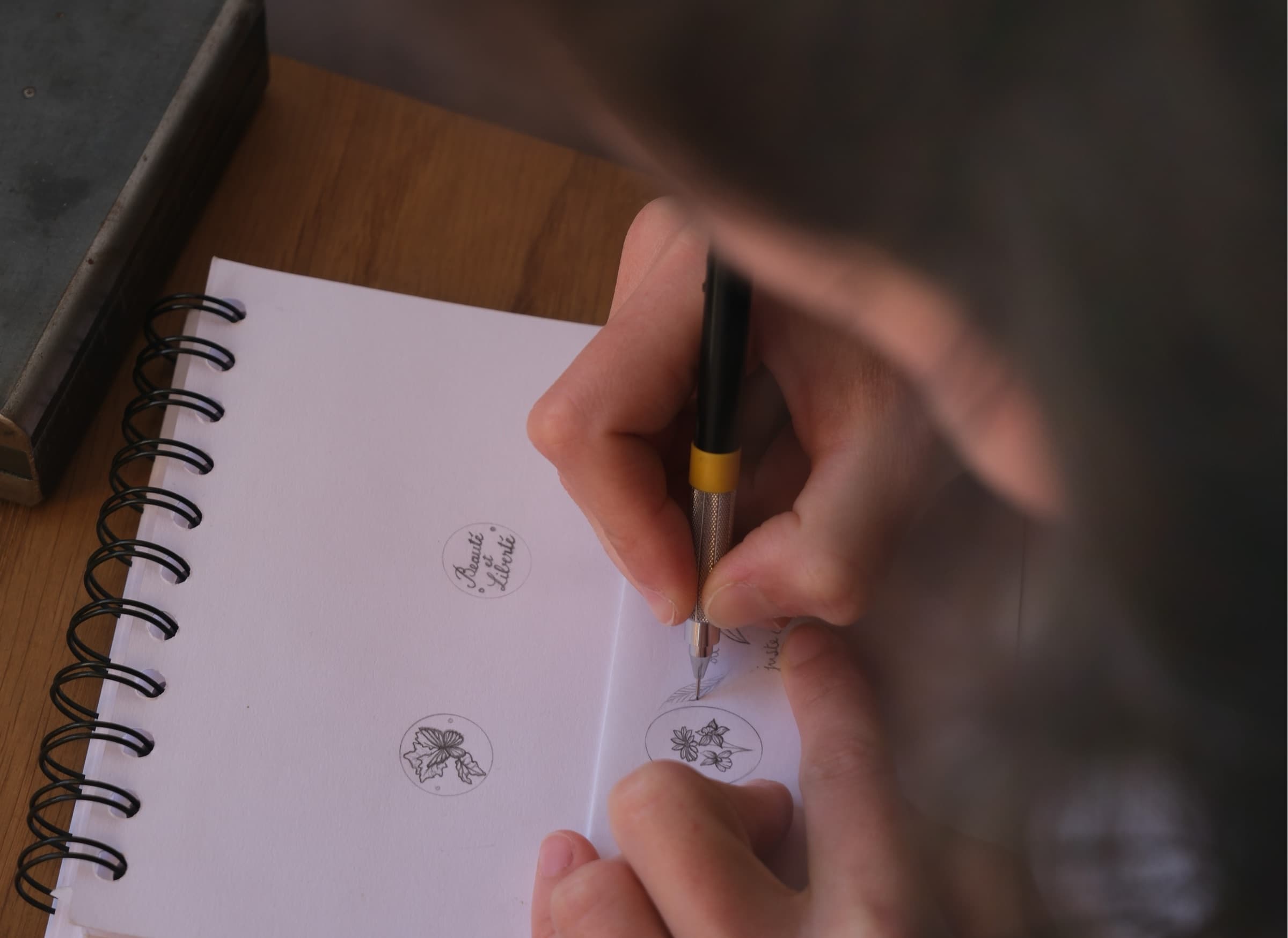 Maëva dessine au crayon des croquis floraux d'un bijou dans son carnet